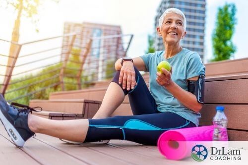 An image of a woman in her sports attire sitting beside a rolled up yoga mat with an apple on her hand