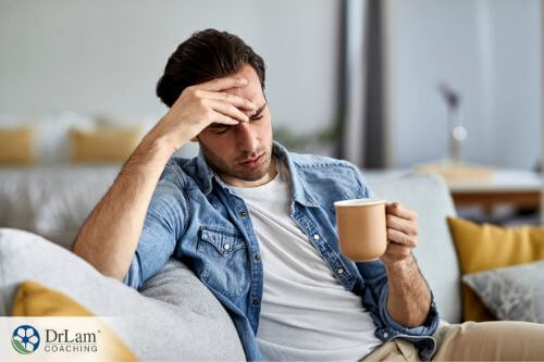 An image of a man sitting on the couch with a cup of coffee and holding his forhead