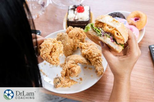 An image of someone eating a burger while having a plate of chicken, donuts, and cake in front of her