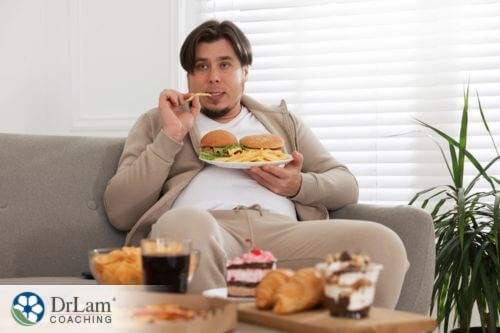 An image of an over weight man sitting on the couch as he eats unhealthy food
