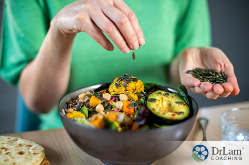 An image of a bowl of cooked veggies and a person sprinkling pumpkin seeds on top