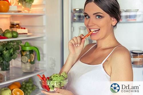 An image of a woman eating vegetables with the refrigerator open