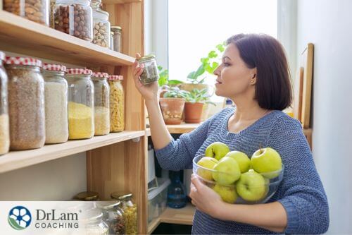 An image of a woman holding a bowl of apples and checking the contents of a jar in her pantry