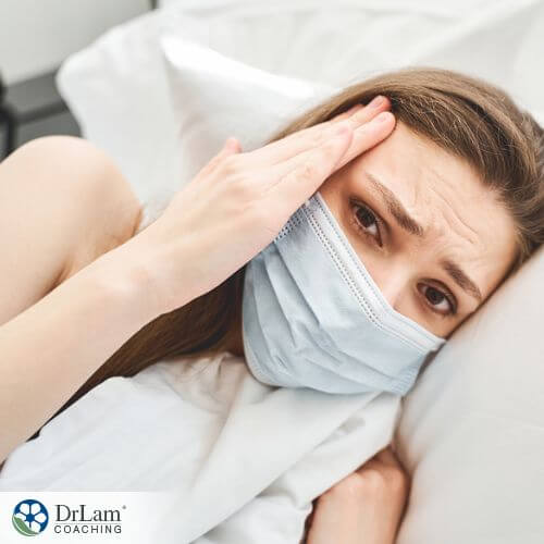 An image of a woman laying in bed wearing a mask and holding her temples