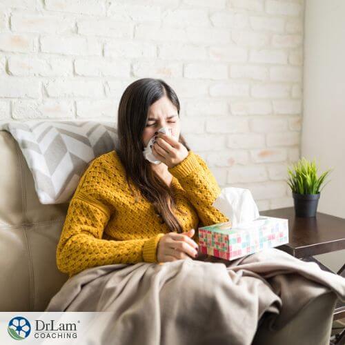 An image of a woman sitting on the couch as she sneezes into some tissue