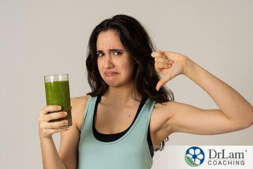 An image of a woman holding a glass of green juice with her thumb pointed down and frowing
