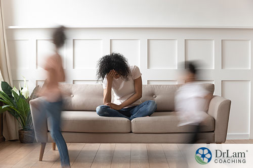 An image of a young mother sitting on the couch holding her head while her children run around