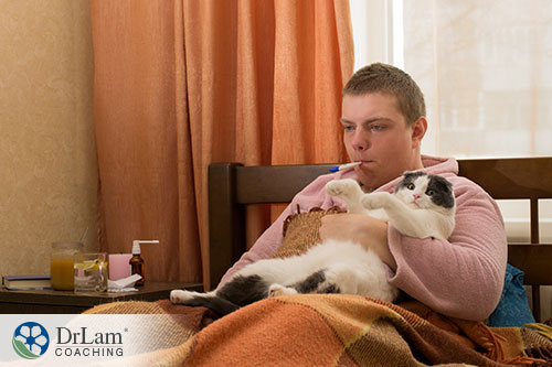 An image of a sick man in bed holding a cat with a thermometer in his mouth.