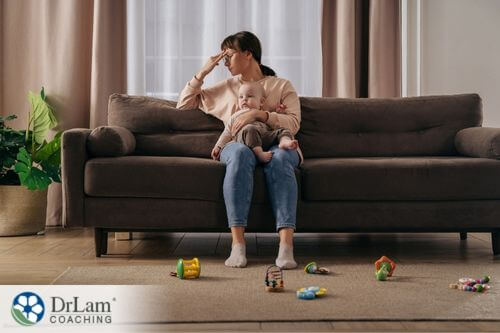 An image of a fatigued mother sitting on the couch as she holds her baby and her forehead