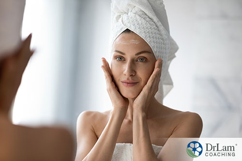 An image of a woman in a white towel looking in the mirror as she's moisturizing her face