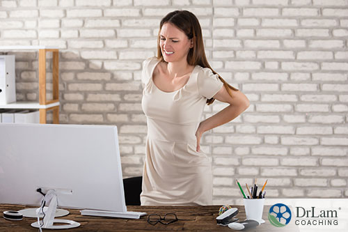 An image of a young woman standing at her desk and holding her back