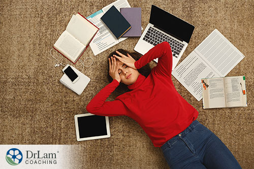 An image of a stressed out young woman laying on the floor surrounded by her laptop, books, notes and cellphone as she holds her head