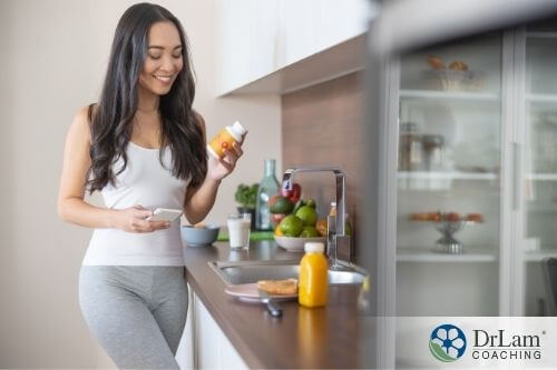 An image of a woman standing in the counter while holding a bottle of supplements in her hand and a breakfast in front of her